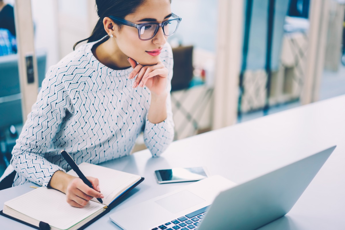 Mujer investigando en su laptop mientras toma notas